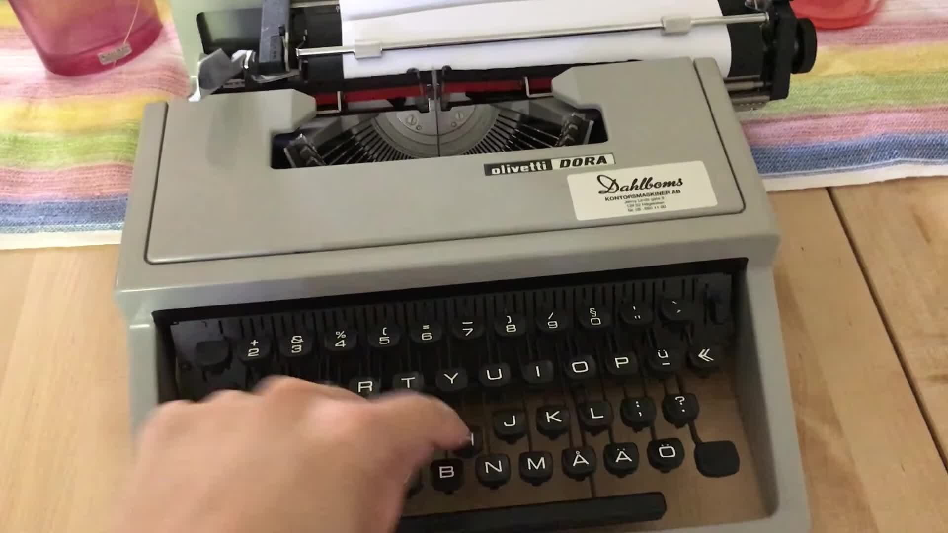 Máquina de escrever Olivetti Dora sobre uma mesa, com uma mão na tecla.